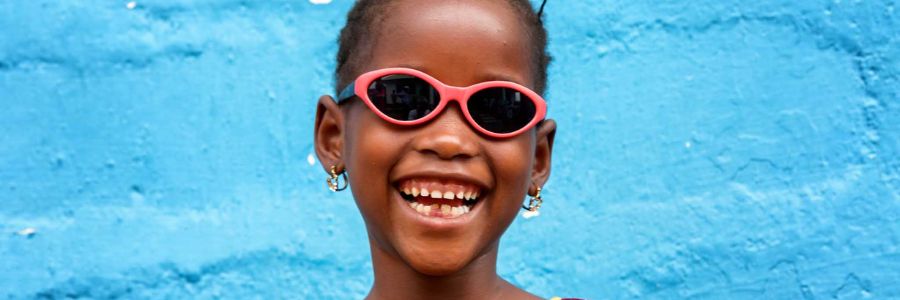 Young girl standing in front of a blue wall in Sierra Leone, Africa wearing pink prescription sunglasses and smiling